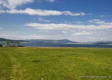 A serene landscape with a grassy field, a lake, and majestic mountains in the background.