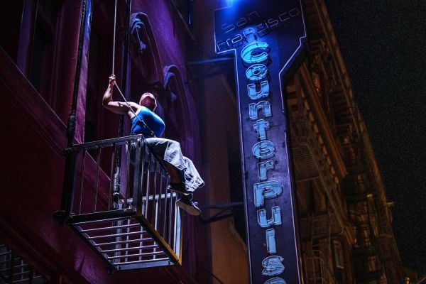 A person with dark skin wearing a blue shirt and gray pants sits on the edge of a large metal cage suspended from the side of a building, hanging from a wire. A blue neon sign reads “San Francisco CounterPulse”