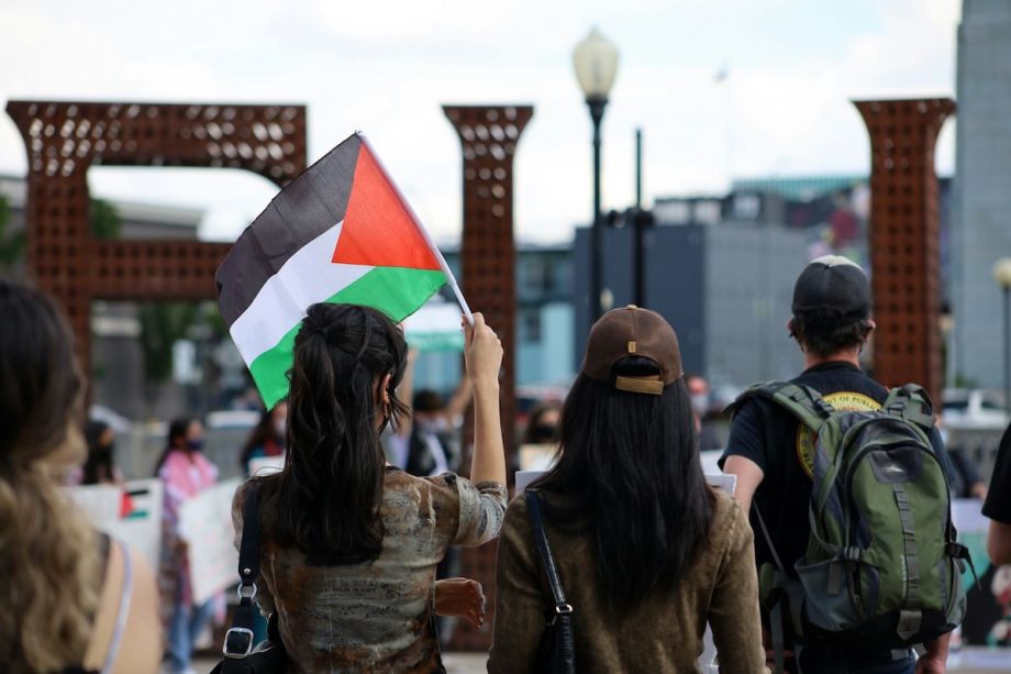 The backs of three people; the person on the left holds up a Palestinian flag.