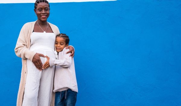 Pregnant person stands in front of a blue wall, with a child hugging their left side